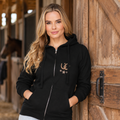 A woman with blonde hair stands in front of a rustic wooden barn door wearing a black zip-up hoodie with a gold embroidered logo. The logo features a horseshoe and stars.