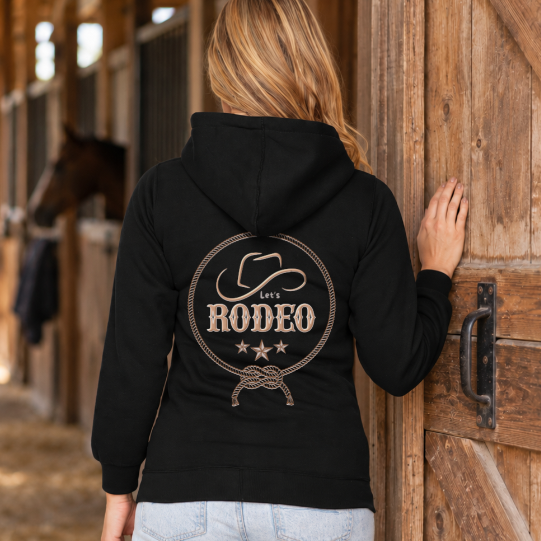 A woman viewed from behind wearing a black hoodie with a large western-themed 'Let's Rodeo' graphic on the back, standing next to a rustic wooden barn door with a horse visible in the background.