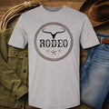 A light gray crewneck t-shirt featuring a black and brown western-style 'RODEO' graphic with a steer skull and rope border, styled in a flat-lay with jeans, a green jacket, a lasso, and a cowboy hat on a wooden background.