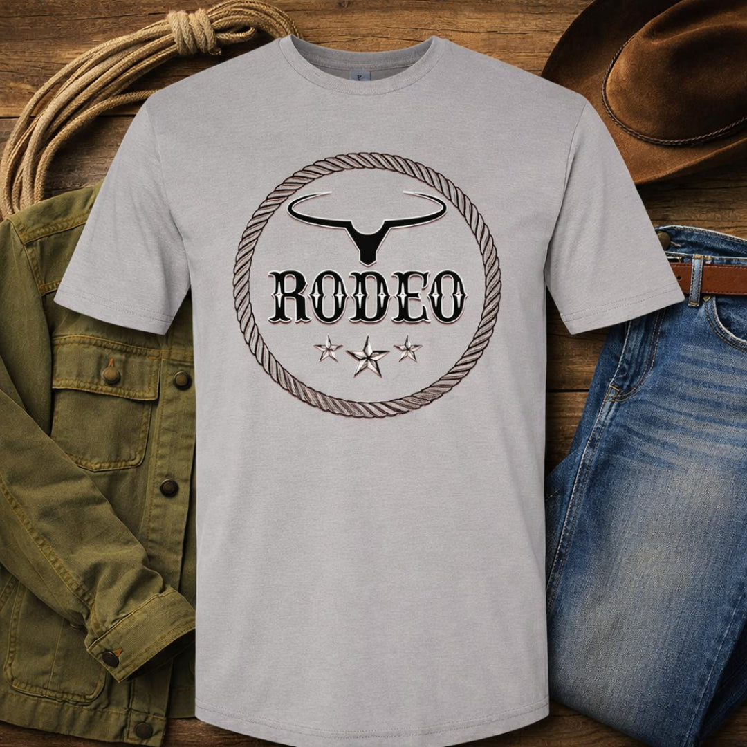 A light gray crewneck t-shirt featuring a black and brown western-style 'RODEO' graphic with a steer skull and rope border, styled in a flat-lay with jeans, a green jacket, a lasso, and a cowboy hat on a wooden background.