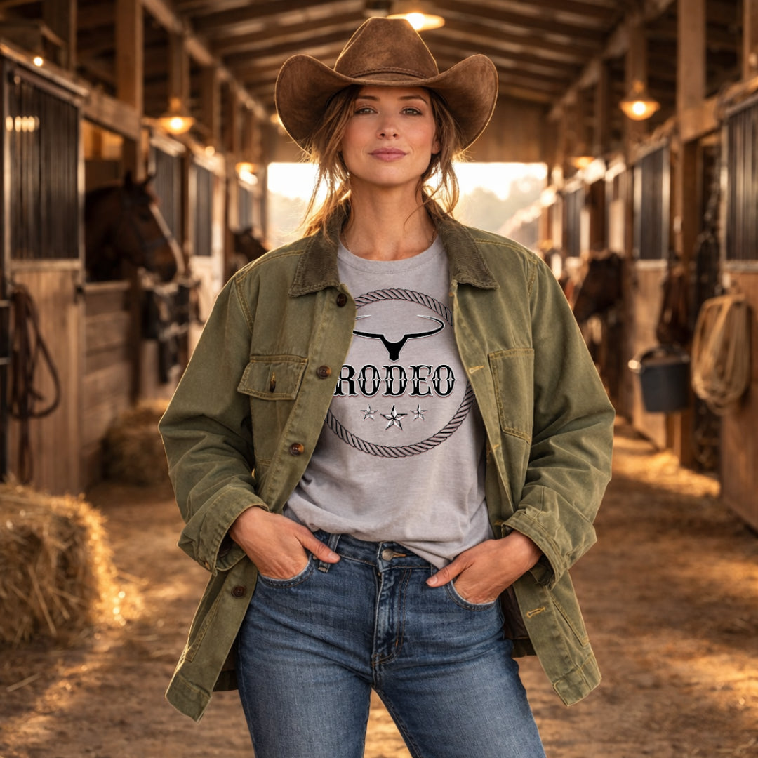 A woman stands in a barn aisle wearing a brown cowboy hat, olive green utility jacket, and a light gray graphic T-shirt with a rodeo bull skull and rope design. Horses are visible in the background stalls.