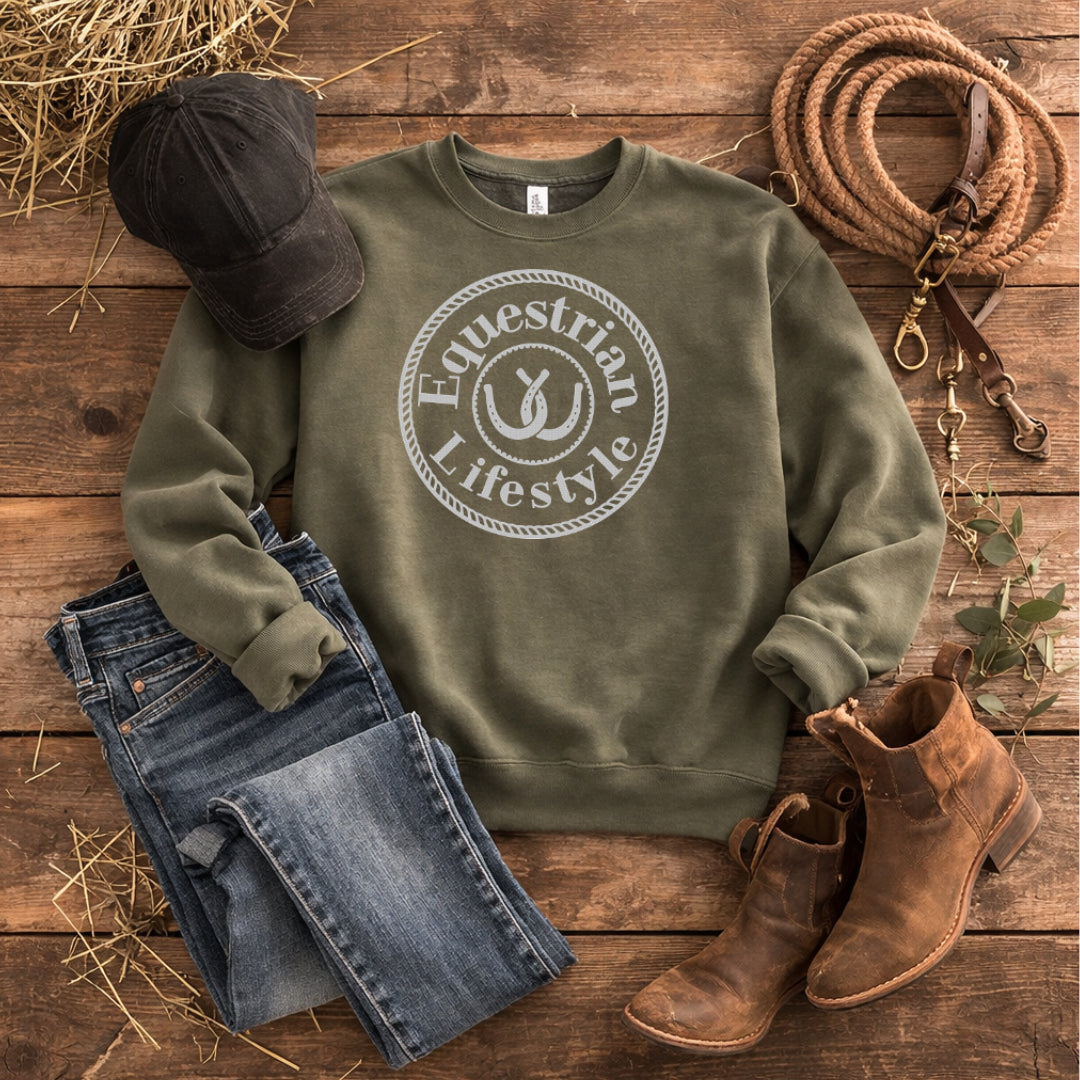 An olive green crewneck sweatshirt with a white circular 'Equestrian Lifestyle' graphic, styled in a flat lay on a rustic wooden background with blue jeans, a black baseball cap, brown leather boots, a rope lasso, and hay.