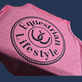 A close-up of a heather pink sleeveless tank top with a large black circular graphic reading 'Equestrian Lifestyle' and a horseshoe emblem, shown against a dark blue background.