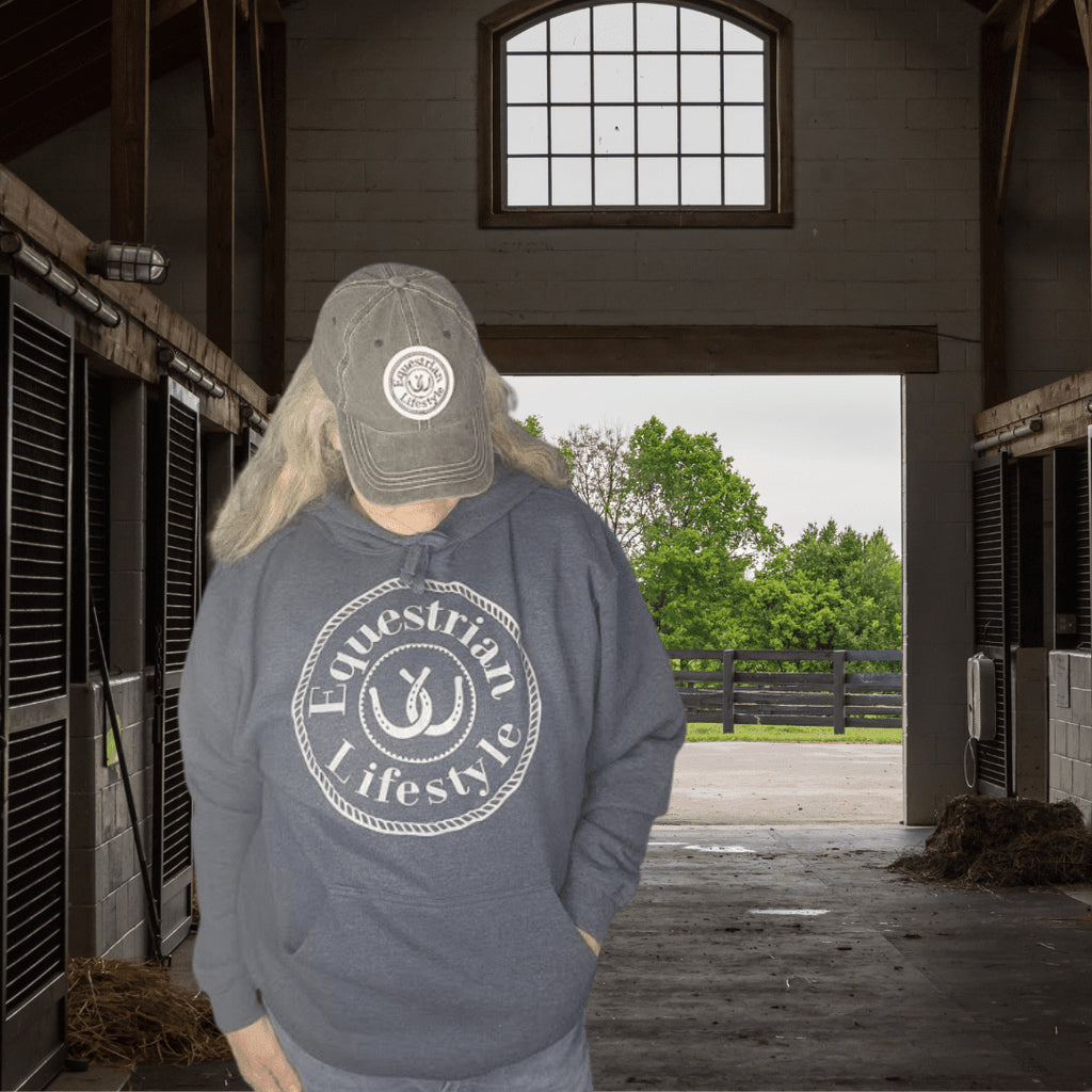A person standing in a barn wearing a gray hoodie with a white circular 'Equestrian Lifestyle' logo on the front, a gray baseball cap, and light-colored pants. The background shows horse stalls on both sides and an open doorway looking out to green trees.