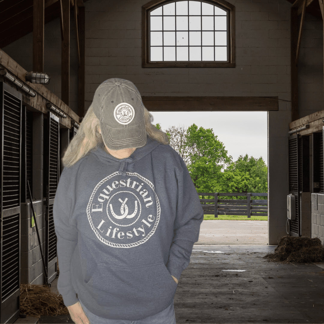 A person standing in a barn wearing a gray hoodie with a white circular 'Equestrian Lifestyle' logo on the front, a gray baseball cap, and light-colored pants. The background shows horse stalls on both sides and an open doorway looking out to green trees.
