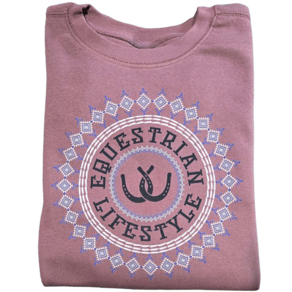 A folded dusty rose t-shirt featuring a circular graphic with the words 'EQUESTRIAN LIFESTYLE' in black, flanked by two black horseshoes, all enclosed in a white and light blue geometric aztec pattern.
