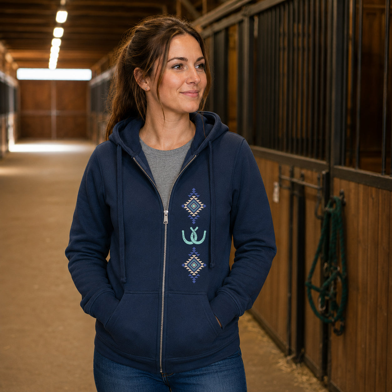 A woman wearing a navy blue zip-up hoodie with a gray crewneck shirt and blue jeans, standing in a horse stable corridor.