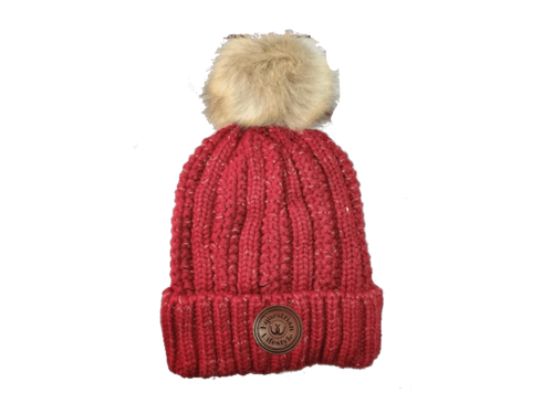 A red ribbed knit beanie hat with a fluffy beige faux-fur pom-pom on top and a circular brown equestrin lifestyle brand patch on the front cuff, shown against a plain white background.