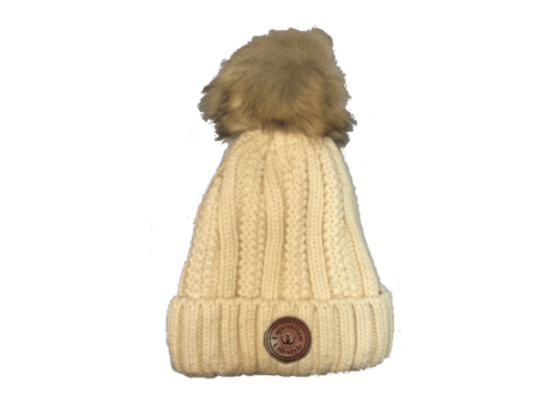 A cream-colored cable-knit beanie hat with a brown faux-fur pom-pom on top and a circular brown leather equestrian lifestyle patch on the front cuff.