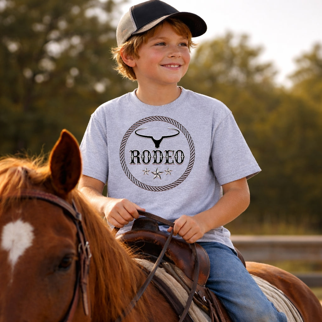 A young boy smiling while riding a brown horse outdoors, wearing a light heather gray t-shirt with a black western-style 'RODEO' graphic on the chest, blue jeans, and a baseball cap.