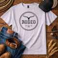 A light heather gray t-shirt with a black and brown rope-style circular graphic that reads 'RODEO' with a steer skull above and stars below. The shirt is styled in a flat lay on a wooden surface with jeans, a leather belt, boots, a baseball cap, sunglasses, rope, and wheat.