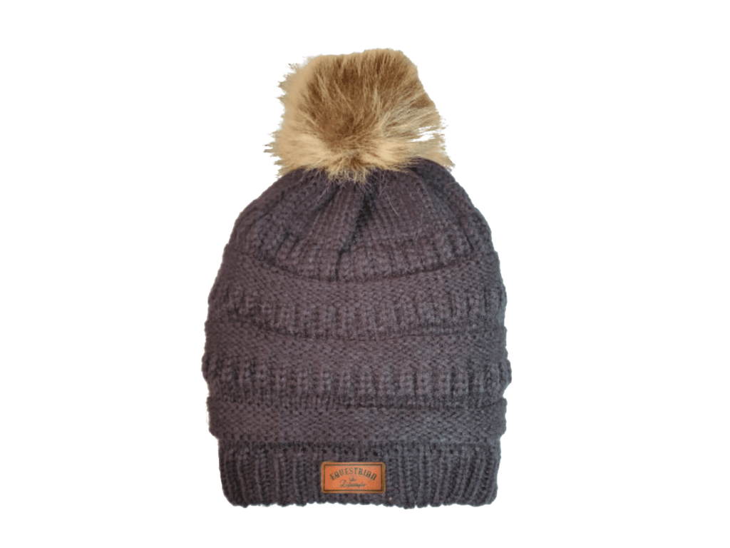 A dark gray ribbed knit beanie hat with a fluffy tan faux-fur pom-pom on top and a small brown leatherette equestrian lifestyle  brand patch on the front cuff.